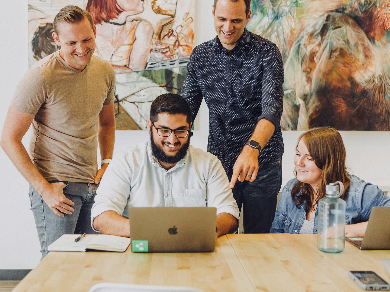 Four colleagues smiling as they gather around a laptop at a shared table